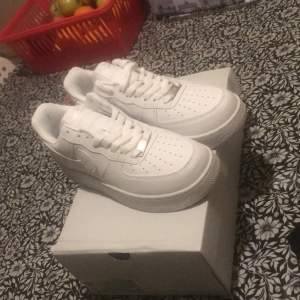 Säljer ett par klassiska vita Nike Air Force sneakers i nytt skick. De är helt vita med snörning och har den ikoniska Nike-loggan på sidan och hälen. Perfekta för både vardag och fest! Skorna kommer i originalkartong. De är 100% äkta 