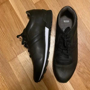 Hugo Boss skor - Snygga svarta skor från Hugo Boss i syntetmaterial. De har en stilren design med snörning och en vit detalj på sulan som ger en modern touch. Perfekta för både vardag och festliga tillfällen. Skorna är i mycket bra skick och redo att användas direkt!