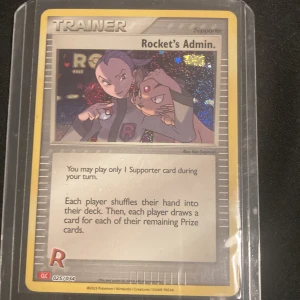 Pokémonkort - Rocket's Admin Charizard Deck Holo TCG - Säljer ett Pokémonkort med titeln 'Rocket's Admin'. Kortet är en tränarkort av typen Supporter och har en holografisk yta. Det är i bra skick och förvaras i en plastficka för extra skydd. Perfekt för samlare eller spelare som vill förstärka sin kortlek.