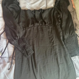 Svart klänning med smockade ärmar - Säljer en elegant svart klänning med smockade långa ärmar och knytning i midjan. Den har en stilren design som passar både till vardags och fest. Klänningen är i bra skick och har en normal passform.
