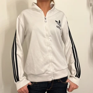 Vit träningsjacka från Adidas - Säljer en snygg vit träningsjacka från Adidas med svarta detaljer. Jackan har dragkedja och långa ärmar med de klassiska tre ränderna längs sidorna. Perfekt för både träning och vardag. Stor Adidas-logga på ryggen för en sportig look.
