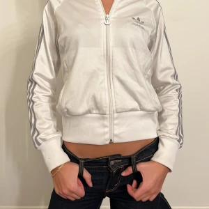 Vit träningsjacka från Adidas - Säljer en snygg vit träningsjacka från Adidas i bra skick. Jackan har dragkedja och långa ärmar med de klassiska tre ränderna längs sidorna. Perfekt för träning eller en sportig vardagslook. Passar bra till både jeans och träningskläder.