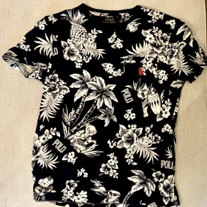 Svart blommig t-shirt från Ralph Lauren - Säljer en snygg svart t-shirt från Ralph Lauren med ett coolt blommönster i vitt. Den har en liten röd logga på bröstet och är i custom slim fit. Perfekt för sommaren eller en avslappnad dag ute. 🌺
