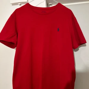 Röd t-shirt från Polo Ralph Lauren - Säljer en klassisk röd t-shirt från Polo Ralph Lauren i bomull. Den har en liten blå logga på bröstet och är perfekt för en avslappnad stil. T-shirten är kortärmad och i mycket bra skick. Perfekt för sommardagar eller som en bas i garderoben.