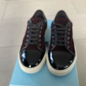 Snygga sneakers med glansig tå - Säljer ett par stiliga sneakers i mycket bra skick. De har en mörkröd färg med glansig svart tå och vita sulor. Perfekta för både vardag och fest! Skorna har snörning och är i en klassisk design som funkar året runt.