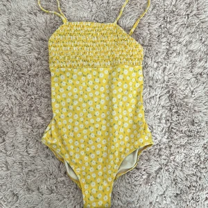 Gul blommig baddräkt - Superfin gul baddräkt med sött blommönster och smala axelband. Den har en smockad överdel som ger en skön och flexibel passform. Perfekt för sommarens alla badäventyr! 🌼