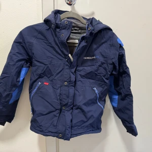 Blå vinterjacka och byxor från Didriksons stl 130 - Säljer ett set med en blå vinterjacka och matchande byxor från Didriksons. Jackan har en avtagbar huva, dragkedja och knappar framtill, samt reflexdetaljer. Byxorna har justerbara hängslen och förstärkta knän. Reflex på högerbyxbeben är borta .Perfekt för kalla vinterdagar! 