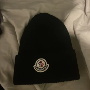 Svart mössa från Moncler - Säljer en snygg svart mössa från Moncler. Den är ribbad och har Monclers ikoniska logga framtill. Perfekt för att hålla sig varm under kalla dagar. Passar till de flesta outfits och är ett måste för vintern! 🖤