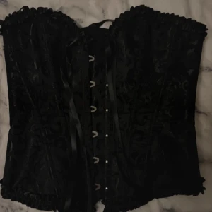 Svart korsett med snörning - Snygg svart korsett med vackert mönster och snörning bak. Den har hakknäppning fram och volangdetaljer upptill och nedtill. Perfekt för en festlig kväll eller en speciell tillställning. Ger en elegant och stilren look.