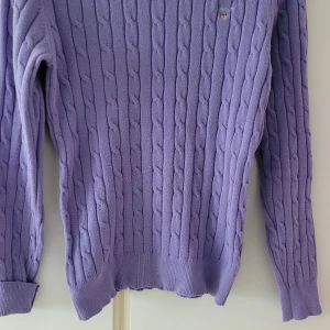 Lila stickad tröja från GANT - Säljer en snygg lila stickad tröja från GANT i stretchig bomull. Perfekt för både höst och vår med sitt kabelstickade mönster. Tröjan har långa ärmar och en klassisk passform. Superfin att matcha med jeans eller kjol!