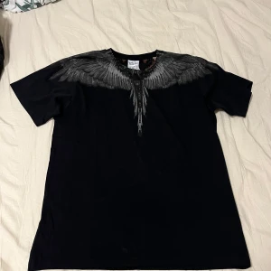 Marcelo Burlon Svart t-shirt - Säljer en svart t-shirt från Marcelo Burlon med ett coolt vingmönster i grått över axlarna. Perfekt för en avslappnad stil eller en utekväll. T-shirten är kortärmad och i mycket bra skick. Passar perfekt till jeans eller shorts!
