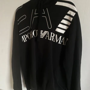 Emporio Armani - Säljer en snygg svart hoodie från Emporio Armani med stort vitt tryck på framsidan. Den har en klassisk design med huva och dragsko. Perfekt för en casual look eller att mysa i hemma. Passar till alla tillfällen!