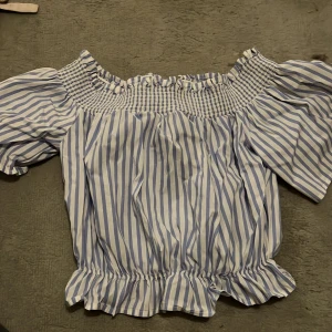 Randig offshoulder-top - Säljer en superfin blå- och vitrandig offshoulder-top med korta puffärmar. Den har en elastisk smockdetalj upptill och vid midjan för en bekväm passform. Perfekt för sommarens alla äventyr! 💙