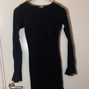 Svart långärmad klänning med volangdetaljer - Snygg kroppsnära svart klänning i mycket bra skick. Den har långa ärmar och fina volangdetaljer vid ärmslut och nederkant. Perfekt för höst och vinter då tyget är tjockt. Passar bra för en elegant och stilren look.