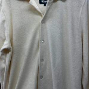 Beige långärmad skjorta från Nithberk - Säljer en stilren beige långärmad skjorta från Nithberk. Skjortan har en klassisk krage och knappar framtill. Perfekt för både vardag och festliga tillfällen. Materialet känns mjukt och bekvämt mot huden. Passar bra till höst och vår.