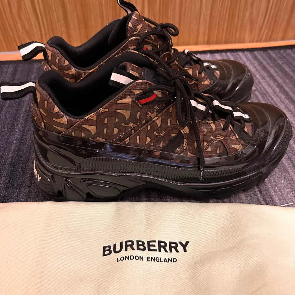 Säljer ett par snygga Burberry sneakers i brunt med coolt mönster och svarta detaljer. De har en robust sula och snörning framtill. Perfekta för att ge din outfit en lyxig touch. Använda men i mycket bra skick. Kommer med originalpåse! Säljer åt en kompis och hon köpte skorna för 9 000 kr. . Skor.