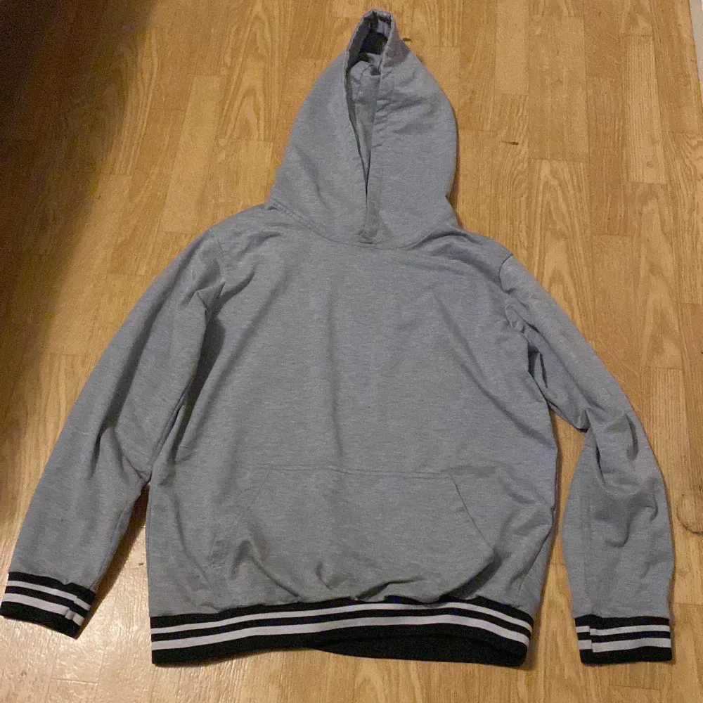 Säljer en grå hoodie i bra skick. Den har en klassisk design med randiga muddar i svart och vitt vid ärmslut och nederkant. Perfekt för en avslappnad stil under höst och vår. Skön och enkel att matcha med det mesta!. Hoodies.