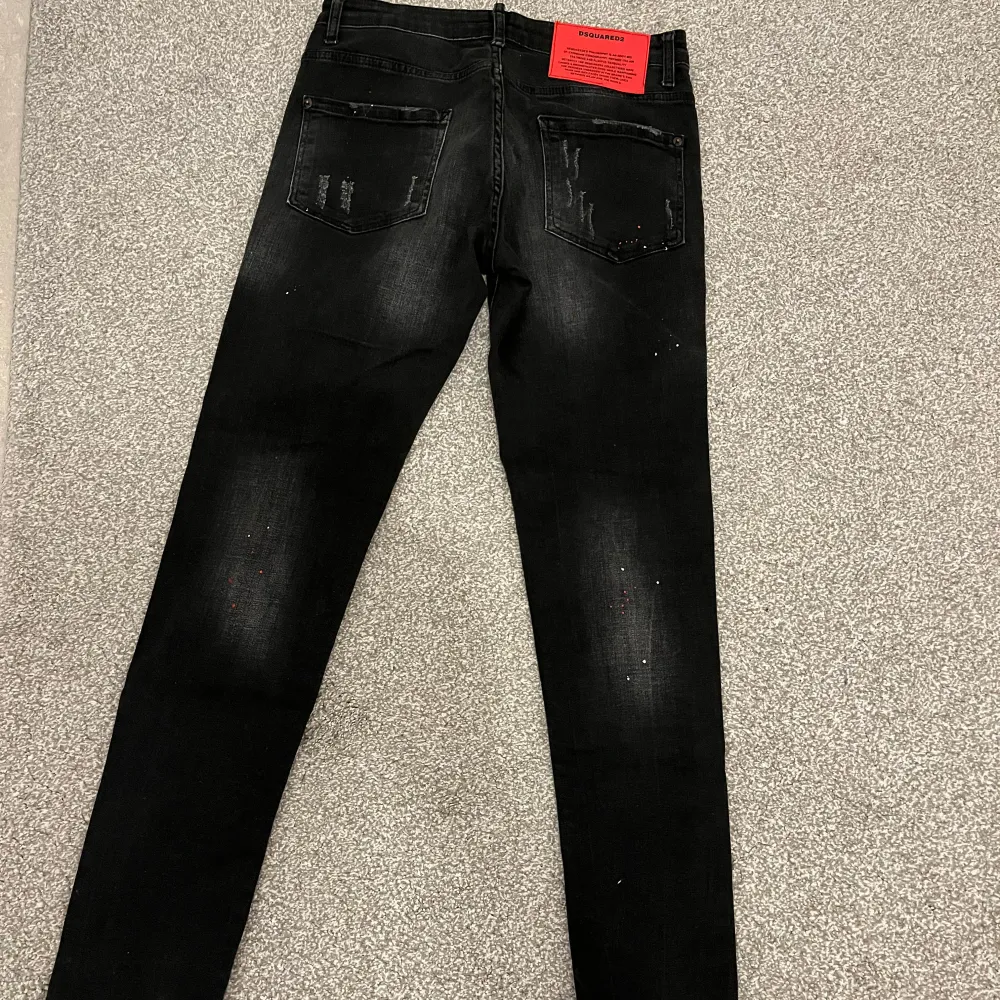 Säljer ett par svarta jeans Dsquared2 liknanade i bra skick. De har en cool, sliten look med ljusare partier och små färgstänk. Jeansen har en slim passform och är perfekta för både vardag och fest. De har knappar i gylfen och klassiska fem fickor.. Jeans & Byxor.