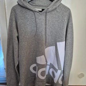 Grå hoodie från Adidas - Säljer en snygg och bekväm grå hoodie från Adidas i storlek XL. Den har en stor vit logga på framsidan och är perfekt för både träning och vardag. Hoodien är i mycket bra skick och har en klassisk passform med långa ärmar och en skön huva. Perfekt för höst och vinter!