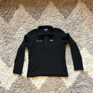 Svart fleece half zip-up tröja, Patagonia - Säljer en svart fleece tröja från Patagonia i storlek L. Den har en dragkedja vid halsen och broderad text 'DeLille Cellars' på bröstet. Perfekt för kyliga dagar och gjord av 100% återvunnen polyester. Supermjuk och bekväm! Women size