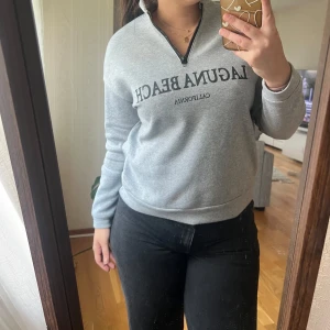 Grå tröja - Säljer en grå sweatshirt med texten 'Laguna Beach California' tryckt på framsidan. Tröjan har en halv dragkedja vid halsen och långa ärmar. Perfekt för en avslappnad stil och passar bra till jeans. Superbekväm och stilren! 