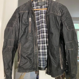 Svart skinnjacka från Jofama - Säljer en svart skinnjacka från Jofama i mycket bra skick. Den har en cool quiltad design på axlarna och dragkedjor både fram och på fickorna. Insidan är fodrad med ett rutigt mönster. Perfekt för höst och vår, och ger en edgy look till vilken outfit som helst!