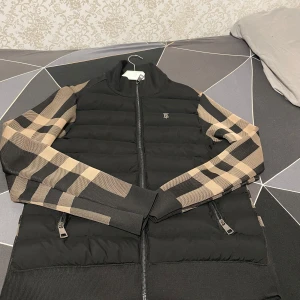 Svart och beige jacka med rutigt mönster - Snygg svart jacka med beige rutiga ärmar från Burberry. Jackan har en dragkedja framtill och ett stilrent märke på bröstet. Perfekt för att hålla stilen i kyligare väder.
