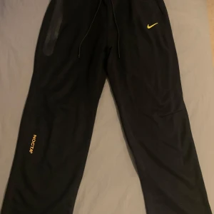 Svarta mjukisbyxor från Nike NOCTA - Svarta mjukisbyxor från Nike NOCTA med gulfärgade detaljer. Byxorna har en dragkedja och snörning i midjan för justerbar passform. Perfekta för en avslappnad stil.