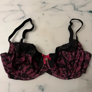 Svart och rosa spets-bh med rosett - Elegant bh i svart och rosa med spetsdetaljer och en söt rosett framtill. Justerbara axelband och en liten charm för extra stil. Perfekt för en speciell kväll.