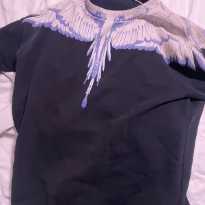 Svart tröja med vingmönster - Säljer en svart tröja från Marcelo Burlon med ett coolt vingmönster i blått och vitt runt halsen. Perfekt för dig som vill ha en unik stil. Tröjan har långa ärmar och är gjord i mjukt material.