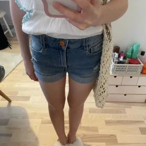 Blå jeansshorts från Kappahl  - Snygga blå jeansshorts från Kappahl med slitna detaljer och uppvikta kanter. Perfekta för en avslappnad stil. De har en knapp och dragkedja framtill samt klassiska fem fickor.