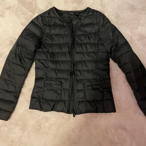 Svart dunjacka - Säljer en stilren svart dunjacka med dragkedja framtill. Den är lätt och smidig att bära, vilket gör den till ett mångsidigt plagg i garderoben. Sen är lite tunnare så lite mer vår eller höst jacka 