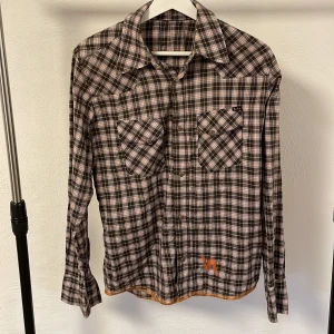  skjorta från Nudie Jeans - Snygg rutig skjorta från Nudie Jeans i brunt, blått och orange. Skjortan har långa ärmar och två bröstfickor med knappar. Den har en broderad detalj i orange vid nederkanten. Perfekt för en avslappnad stil. Pris kan såklart diskuteras!