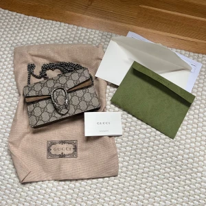 Gucci väska - Säljer en snygg beige axelväska från Gucci med det klassiska GG-mönstret. Väskan har en kedjerem och ett dekorativt spänne framtill. Dionysus GG super mini bag nypris ligger på 11 100 kr. Är som helt ny! 