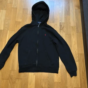 Svart hoodie med dragkedja från Ralph Lauren - Säljer en stilren svart hoodie från Ralph Lauren med dragkedja och huva. Den har en liten röd logotyp på bröstet och praktiska fickor framtill. Perfekt för en avslappnad look.