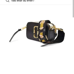 marc jacobs väska - Snygg svart axelväska med en bred, mönstrad rem i gult och rosa. Väskan har en dragkedja och gulddetaljer som ger en lyxig känsla. Perfekt för att bära dina nödvändigheter med stil.