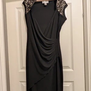 Svart klänning med pärldetaljer - Elegant svart klänning från Adrianna Papell med draperad framsida och pärldetaljer på axlarna. Perfekt för en festlig kväll. 