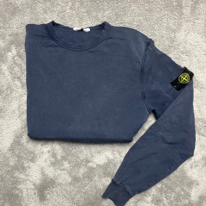 Blå tröja från Stone Island - Säljer en snygg blå tröja från Stone Island med deras ikoniska märke på ärmen. Tröjan har långa ärmar och en rund halsringning. Perfekt för en stilren look.
