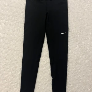 Svarta tights från Nike - Säljer ett par svarta tights från Nike med en liten vit logga på benet. De är designade för en tight passform och är perfekta för träning. Materialet är syntetiskt och ger bra stöd och komfort.