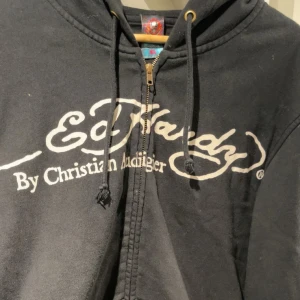 Svart hoodie från Ed Hardy - Säljer en svart hoodie från Ed Hardy med dragkedja och stort tryck av en dödskalle med hatt och kortlek. Hoodien har justerbar huva med snören och ribbade muddar. 
