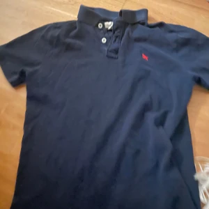 Mörkblå pikétröja med röd logga - Snygg mörkblå pikétröja med klassisk krage och knappar. Den har en liten röd logga på bröstet som ger en stilren touch. Perfekt för en avslappnad men ändå uppklädd look.