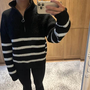 Svart och vit randig stickad tröja med dragkedja - Säljer en mysig svart och vit randig stickad tröja med dragkedja i halsen. Perfekt för kyliga dagar med sin bekväma passform och stilrena design.