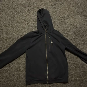 Svart hoodie från Sail Racing - Säljer en svart hoodie från Sail Racing med dragkedja och huva. Den har en mjuk insida och en liten logotyp på bröstet. Tröjan passar främst xs