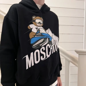 Svart hoodie från Moschino med björntryck - Säljer en svart hoodie från Moschino med ett stort tryck av en björn på framsidan. Hoodien har en klassisk passform med långa ärmar och en bekväm huva. Perfekt för en avslappnad stil.