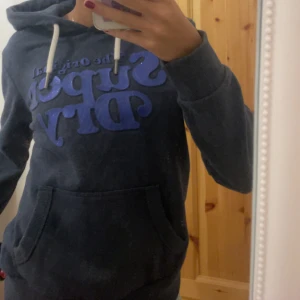 Mörkblå hoodie från Superdry - Säljer en snygg mörkblå hoodie från Superdry med blå text på framsidan. Den har en klassisk känguruficka och justerbar huva med vita snören. Perfekt för en avslappnad stil.