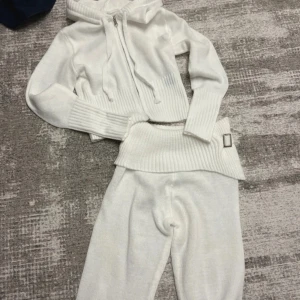 Vit stickad hoodie med dragkedja och mjukisbyxor - Säljer en vit stickad hoodie med dragkedja och ribbade detaljer. Tröjan har långa ärmar och en mysig huva med knytband. Perfekt för kyliga dagar!