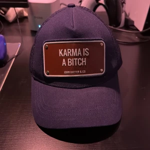 Mörkblå keps från John Hatter & Co - Snygg mörkblå keps med texten 'Karma is a bitch' på en metallplatta framtill. Kepsen har en klassisk design och är perfekt för en avslappnad stil. Märket John Hatter & Co är broderat på insidan.