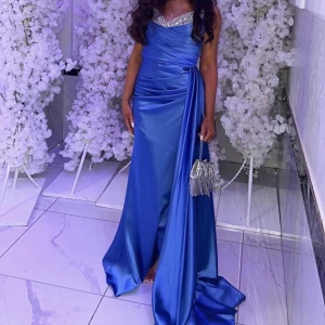 Blå långklänning med glittrig detalj som är egen designad. Storlek xs-s. Pris: 3000 kr kan diskuteras - Elegant blå långklänning med en glittrig överdel och draperad design. Klänningen har en slits och en vacker drapering som ger en lyxig känsla. Perfekt för speciella tillfällen.