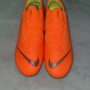 Orange fotbollsskor från Nike - Snygga och iögonfallande orange fotbollsskor från Nike med svarta detaljer och snörning. Skorna har en slitstark sula med dobbar för optimalt grepp på planen. Perfekta för den som vill sticka ut på fotbollsplanen. Priset är inte hugget i sten så tveka inte vid minsta fundering.