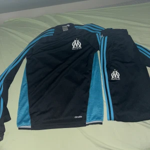 Marseille tracksuit  - Säljer ett svart och blått träningsset från Adidas med Olympique de Marseille-logga. Tröjan har långa ärmar och blå ränder längs ärmarna. Perfekt för fotbollsträning eller bara vanlig wear 